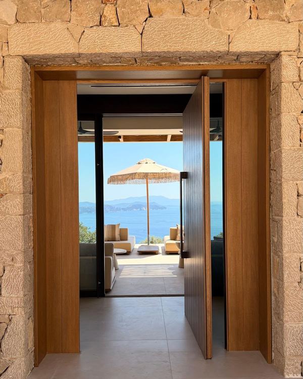 Stone entrance with a tall timber door standing ajar, framing a view straight through the villa to a parasol and the sea.
