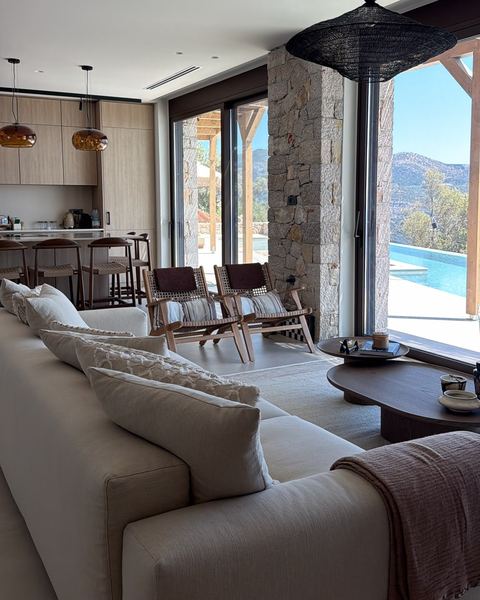 Open-plan living room with a cream sofa, woven armchairs and a stone column, the kitchen behind and the pool outside.