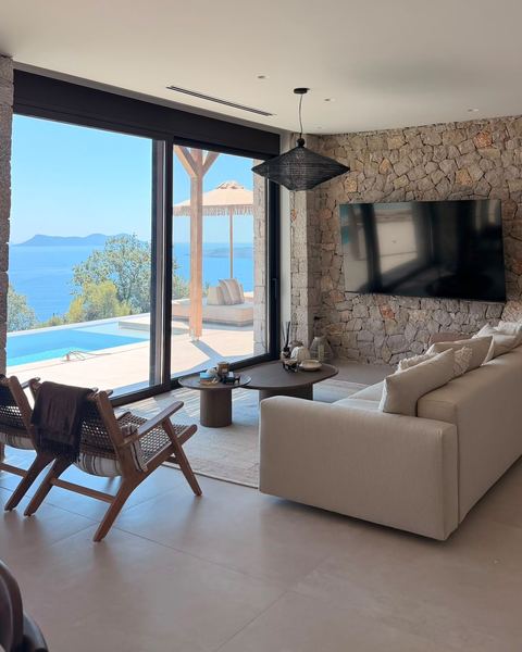 Living room with a cream sofa, a wall-mounted TV on a stone wall and sliding glass doors to the pool and sea.