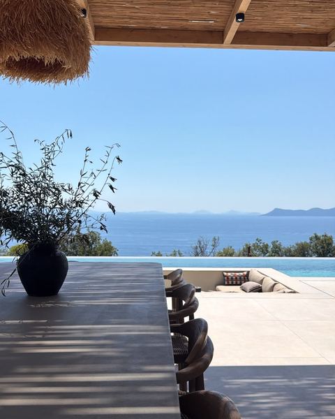 Shaded outdoor dining table with carved wooden chairs under a reed pergola, looking across the pool to the sea.
