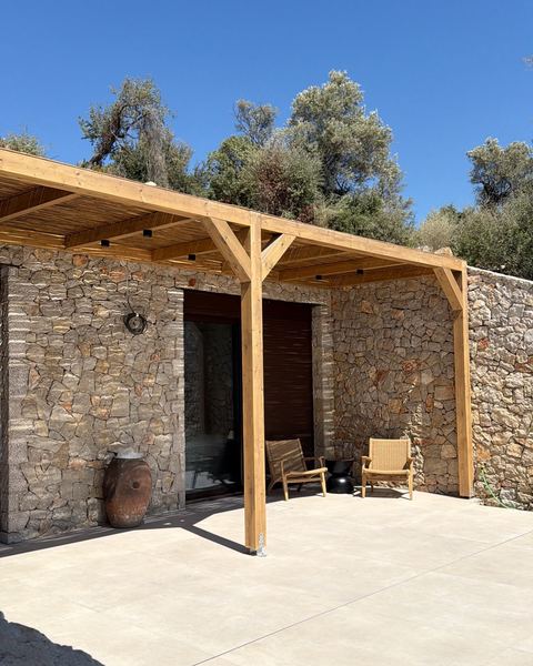 Stone-walled wing with a timber pergola and two woven armchairs, olive trees behind.