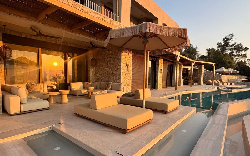 Two-storey stone villa at sunset with a covered lounge, daybeds under a fringed parasol and the pool terrace.